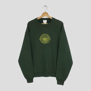 Könnte beinhalten: Dunkelgrünes Sweatshirt mit Rundhalsausschnitt und langen Ärmeln. Auf der Vorderseite befindet sich ein gelbes kreisförmiges Grafikdesign mit dem Text "BATTLE PASS UNIVERSITY" und den Jahreszahlen "1969" und "2013". Das Sweatshirt hängt an einem Holzkleiderbügel.