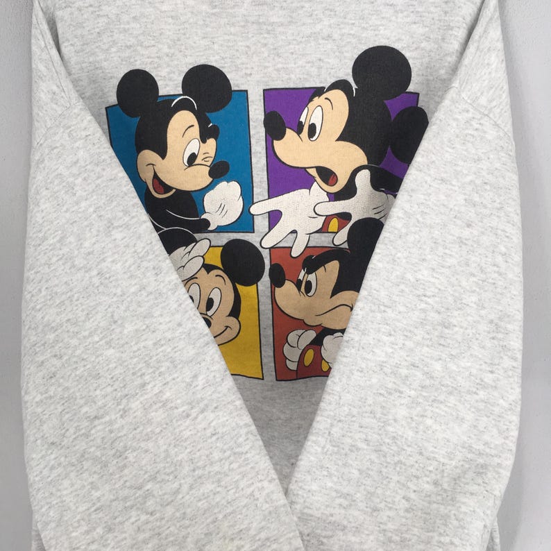 May include: A gray sweatshirt featuring a colorful graphic of Mickey Mouse in four different poses. Each Mickey Mouse is set against a different colored square: blue, purple, yellow, and red. The sweatshirt is folded.