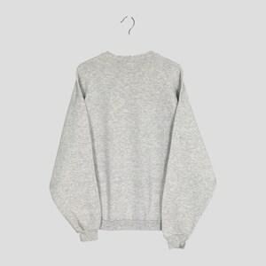 May include: A heather gray sweatshirt hanging from a metal hanger. The sweatshirt has a crew neck, long sleeves, and ribbed cuffs and hem. The fabric appears to be a soft, comfortable material. The background is a solid, neutral color.
