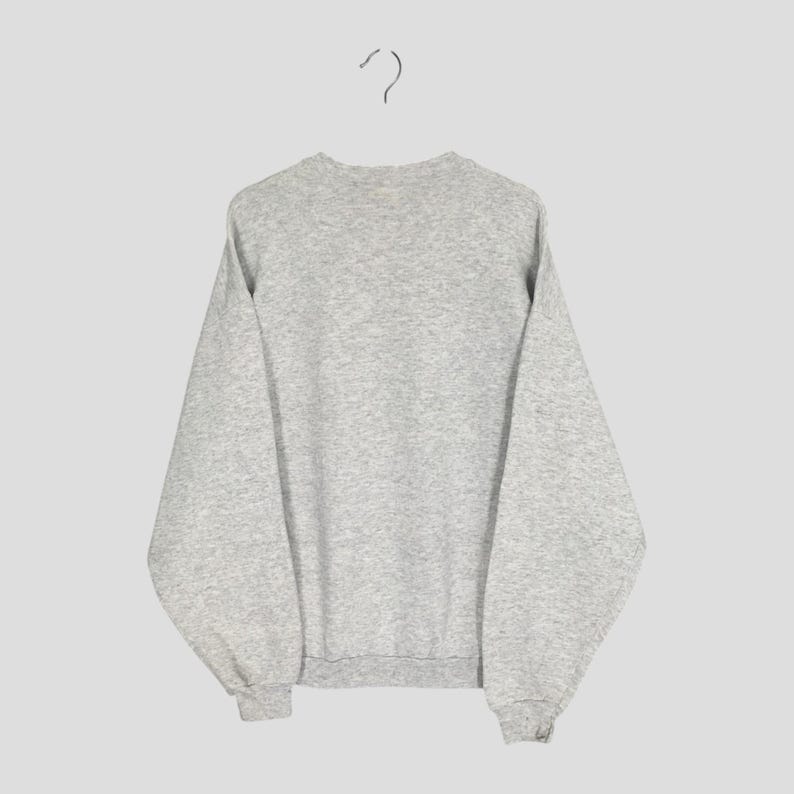 May include: A heather gray sweatshirt hanging from a metal hanger. The sweatshirt has a crew neck, long sleeves, and ribbed cuffs and hem. The fabric appears to be a soft, comfortable material. The background is a solid, neutral color.