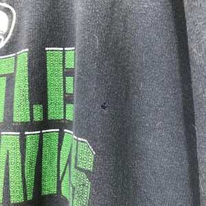 Puede incluir: Primer plano de una camiseta negra con un gr&aacute;fico verde y blanco que dice "Little Kings". La camiseta tiene un peque&ntilde;o agujero en la tela.