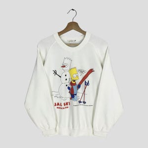 Puede incluir: Sudadera blanca con una imagen de dibujos animados de Bart Simpson esquiando con un muñeco de nieve. El gráfico incluye el texto "JAL SKI HOKKAIDO". El diseño es en rojo, azul, amarillo y negro.