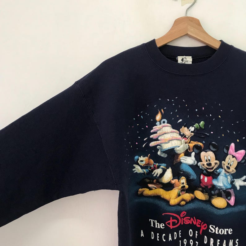 May include: A navy blue sweatshirt with a graphic print of Mickey Mouse, Minnie Mouse, Goofy, Donald Duck, and Pluto celebrating with a birthday cake. The text "The Disney Store A Decade of Dreams 1997" is printed on the bottom of the sweatshirt.