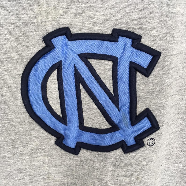 K&ouml;nnte beinhalten: Nahaufnahme eines grauen Stoffes mit einem hellblau und marineblau gestickten Logo. Das Logo sind die ineinandergreifenden Buchstaben "NC" mit einem eingetragenen Warenzeichen.