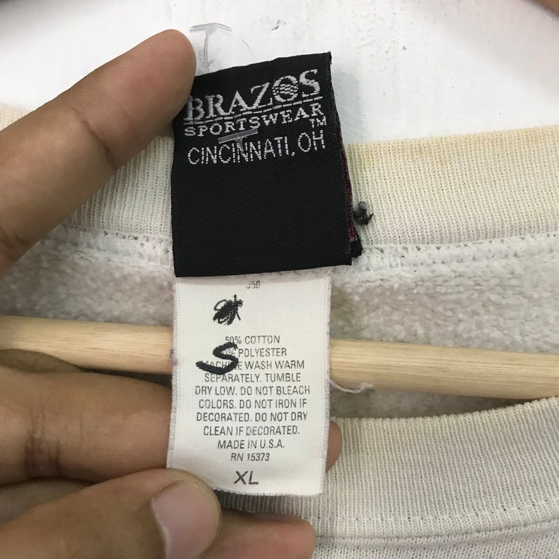 May include: A white sweatshirt with a black tag that reads "Brazos Sportswear TM Cincinnati, OH". The tag also includes care instructions for the sweatshirt, which is made of 50% cotton and 50% polyester. The sweatshirt is size XL.