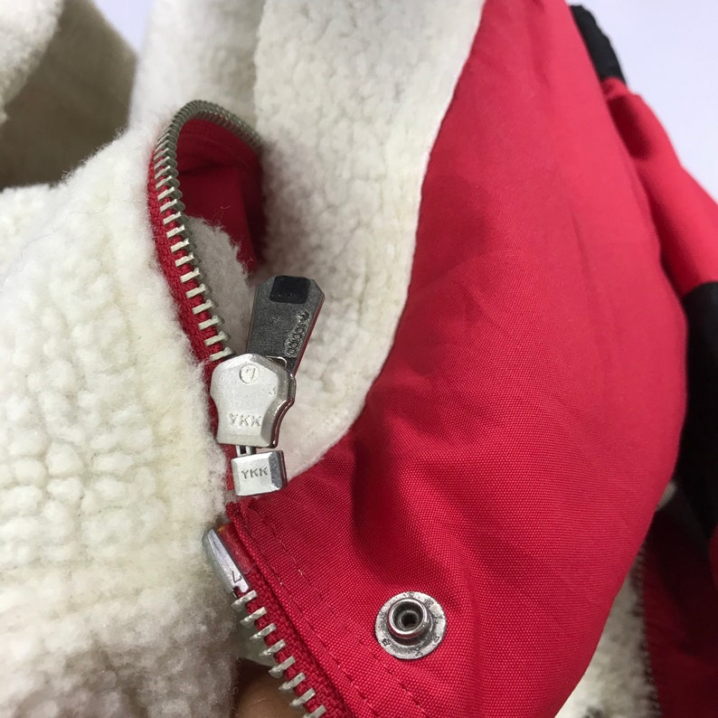 May include: Close-up of a red zipper on a jacket with a white fleece lining. The zipper has a silver pull with the text "YKK" on it.