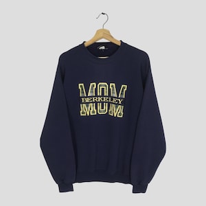 Może przedstawiać: Granatowa bluza z napisem "MUM BERKELEY MUM" w żółtych literach. Litery mają wzór w kratę. Bluza wisi na drewnianym wieszaku.