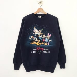 May include: A navy blue sweatshirt with a graphic of Mickey Mouse and friends celebrating. The text "The Disney Store A Decade of Dreams 1997" is printed below the graphic.