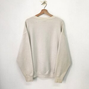 May include: A plain white crew neck sweatshirt with long sleeves. The sweatshirt is hanging on a wooden hanger against a white wall.