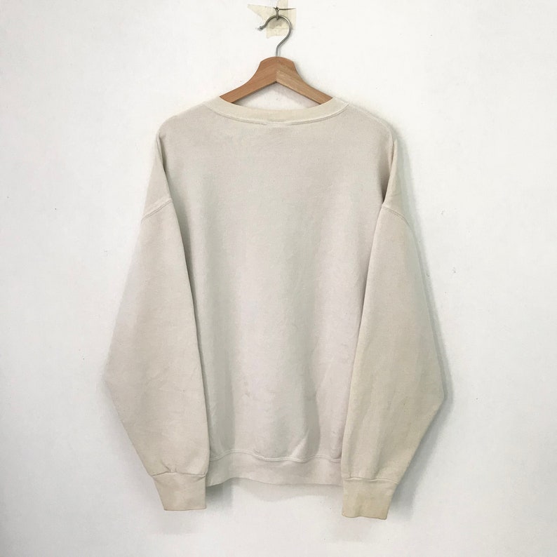 May include: A plain white crew neck sweatshirt with long sleeves. The sweatshirt is hanging on a wooden hanger against a white wall.