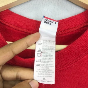 May include: Red long-sleeve shirt with a white tag that says "PEANUTS QUFS" and includes care instructions and size information. The tag also says "50% Cotton/Algodon 50% Polyester/Poliester Made in Honduras Hecho en Honduras XL UPC: 75831535501"
