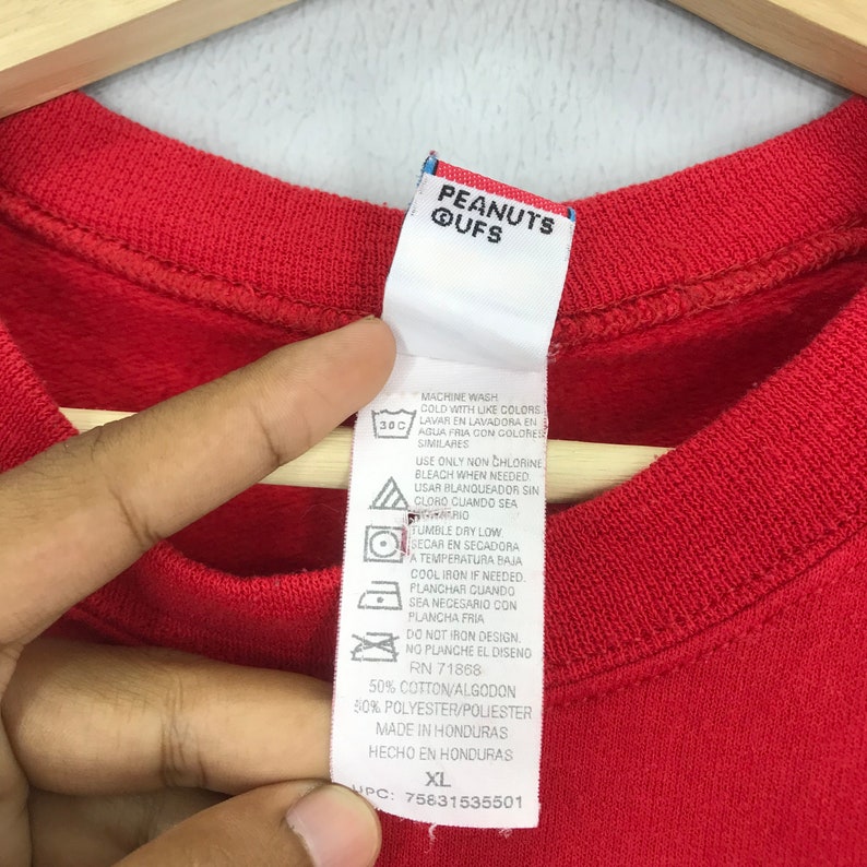 May include: Red long-sleeve shirt with a white tag that says "PEANUTS QUFS" and includes care instructions and size information. The tag also says "50% Cotton/Algodon 50% Polyester/Poliester Made in Honduras Hecho en Honduras XL UPC: 75831535501"