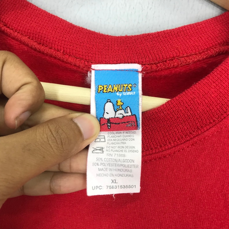 May include: Red Peanuts by Schulz t-shirt with a white tag. The tag has a cartoon image of Snoopy and Woodstock. The tag also includes care instructions and size information. The tag says "Cool iron if needed. Planchar cuando sea necesario con plancha fria. Do not iron design. No planche el diseno. RN 71868. 50% Cotton/Algodon 50% Polyester/Poliester. Made in Honduras. Hecho en Honduras. XL. UPC: 75631535501"