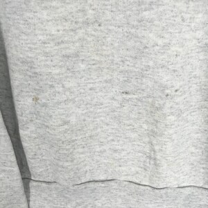 May include: Close-up of a light gray sweatshirt with a ribbed hem. The fabric has a speckled texture and some small dark spots. The sleeve is visible on the left side of the frame.