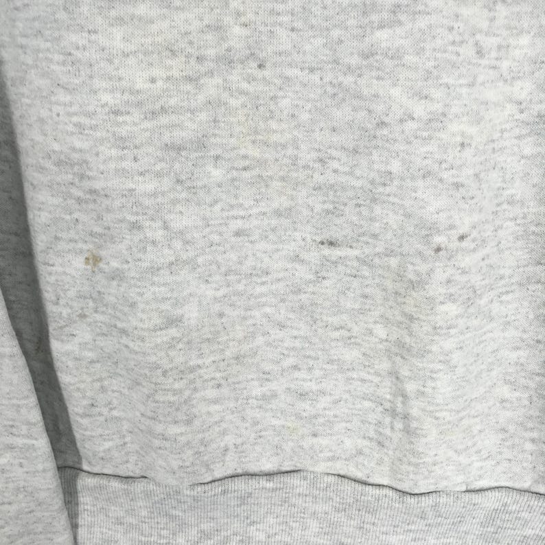 May include: Close-up of a light gray sweatshirt with a ribbed hem. The fabric has a speckled texture and some small dark spots. The sleeve is visible on the left side of the frame.