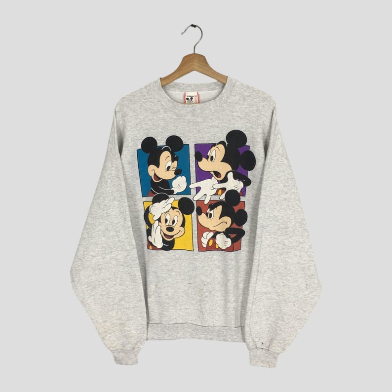 May include: A light gray sweatshirt featuring a four-panel graphic of Mickey Mouse in various poses. Each panel has a different colored background: blue, purple, yellow, and red. The sweatshirt has long sleeves and a crew neck.