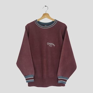 May include: A maroon Champion sweatshirt with a crew neck and long sleeves. The collar and cuffs have a striped design in teal, white, and maroon. The Champion logo is embroidered on the chest in white.
