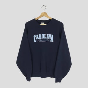 May include: Navy blue sweatshirt with the word "CAROLINA" in light blue arched across the chest, with "TAR HEELS" below. The sweatshirt is hanging on a wooden hanger against a neutral background.