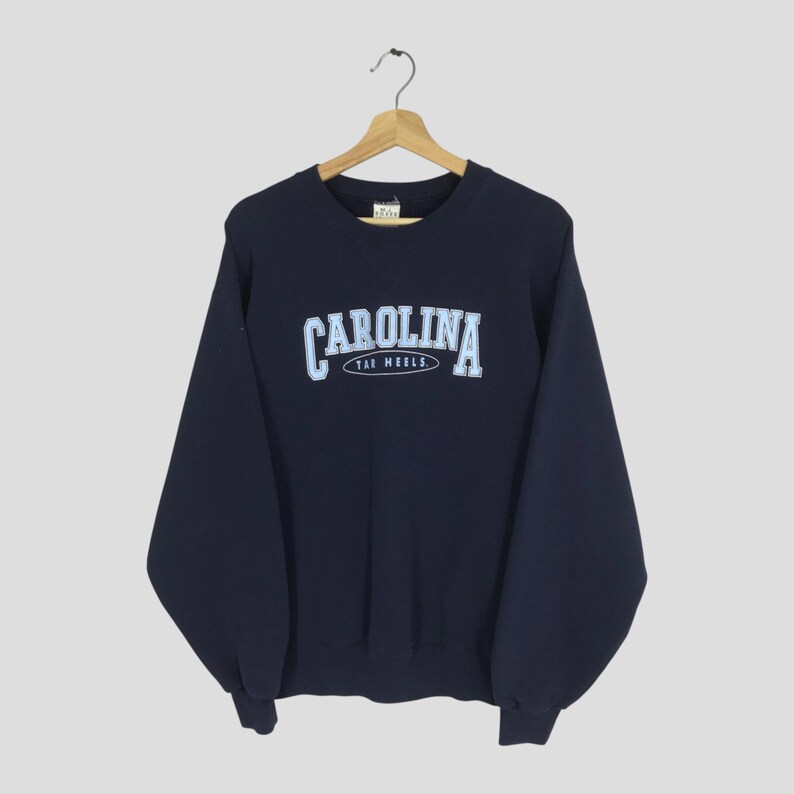 May include: Navy blue sweatshirt with the word "CAROLINA" in light blue arched across the chest, with "TAR HEELS" below. The sweatshirt is hanging on a wooden hanger against a neutral background.