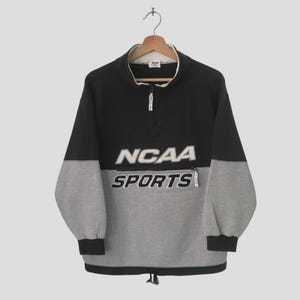 Puede incluir: Un jersey de vellón negro y gris con cierre de cremallera a la mitad. El jersey tiene un gran logotipo de "NCAA SPORTS" en la parte delantera.