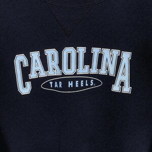 May include: Navy blue sweatshirt with the word "CAROLINA" in light blue block letters with a white outline. Below the word "CAROLINA" is the text "TAR HEELS" in a white oval.