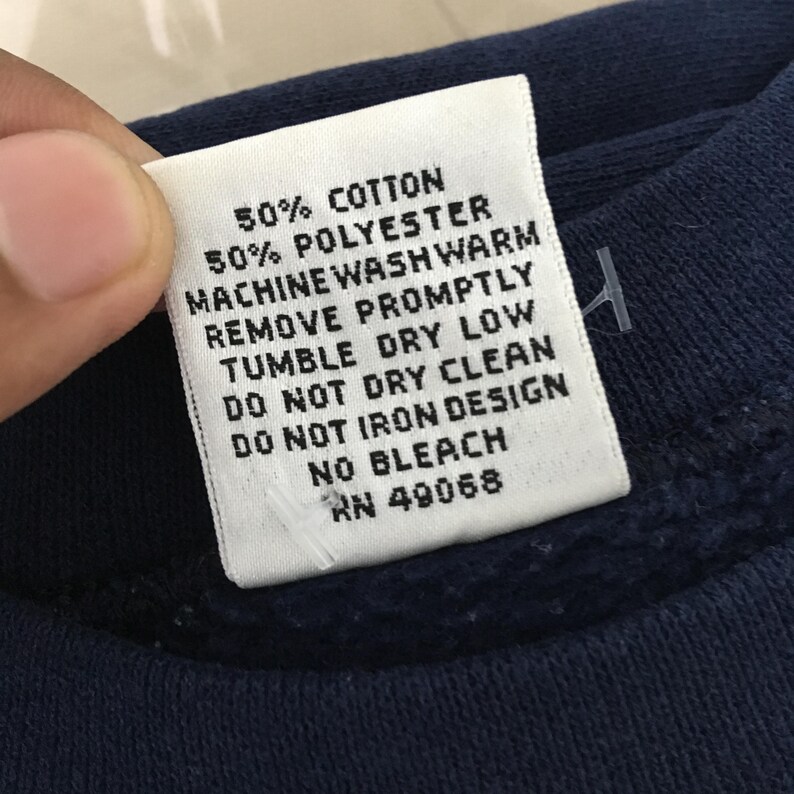 May include: Close-up of a white clothing tag with black text. The tag reads "50% COTTON, 50% POLYESTER, MACHINE WASH WARM, REMOVE PROMPTLY, TUMBLE DRY LOW, DO NOT DRY CLEAN, DO NOT IRON DESIGN, NO BLEACH, RN 49068."
