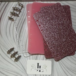 May include: A set of three pink planner inserts, one with a glitter finish, and two with a solid pink finish. The inserts are designed for a six-ring planner and have holes punched along the side. The inserts are displayed on a clear acrylic sheet with a silver six-ring binder.