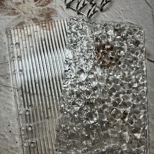 May include: Two clear plastic sheets with different textures. One sheet has a smooth, striped pattern, while the other has a bumpy, bubble-like texture. Both sheets have holes for a binder.
