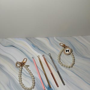 May include: Five gold and pink tools with pearl-embellished handles. The tools have a loop at the top for hanging. The tools are arranged on a white marble surface.