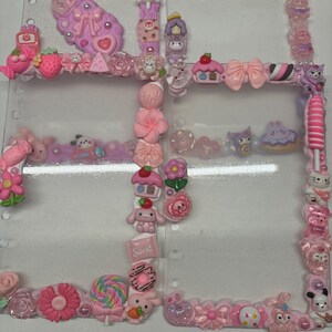 May include: A clear, plastic binder decorated with pink and purple embellishments. The decorations include bows, flowers, cupcakes, and cartoon characters. The binder has a variety of shapes and sizes of charms, creating a whimsical and playful aesthetic.