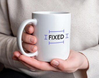 May include: White ceramic mug with the text "I FIXED" in purple. The mug has a curved handle and is held by a person wearing a light-colored sweater. The mug is a standard size for coffee or tea.