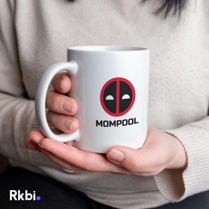 May include: White ceramic mug with a black and red graphic design and the word "MOMPOOL" in black text. The mug has a large handle and is held by a person.