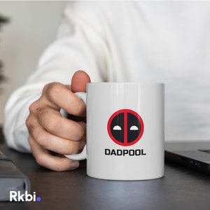 Può includere: Tazza da caffè in ceramica bianca con un cerchio rosso e un logo Deadpool nero. Il testo "DADPOOL" è stampato sotto il logo.