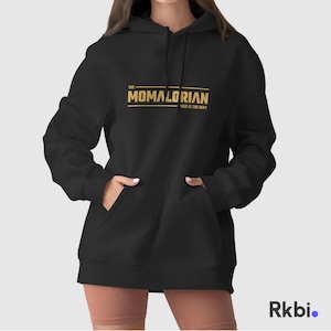 Pu&ograve; includere: Felpa con cappuccio nera con una grafica dorata che dice "The Momalorian This is the Way"