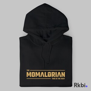 Pu&ograve; includere: Felpa con cappuccio nera con una grafica dorata che recita "THE MOMALORIAN THIS IS THE WAY".