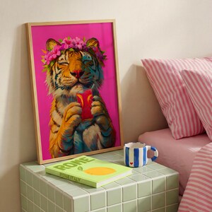May include: A framed wall art print featuring a tiger with closed eyes wearing a pink flower crown and holding a red drink. The tiger has orange and black stripes. The background is a solid bright pink color. The frame is light wood.