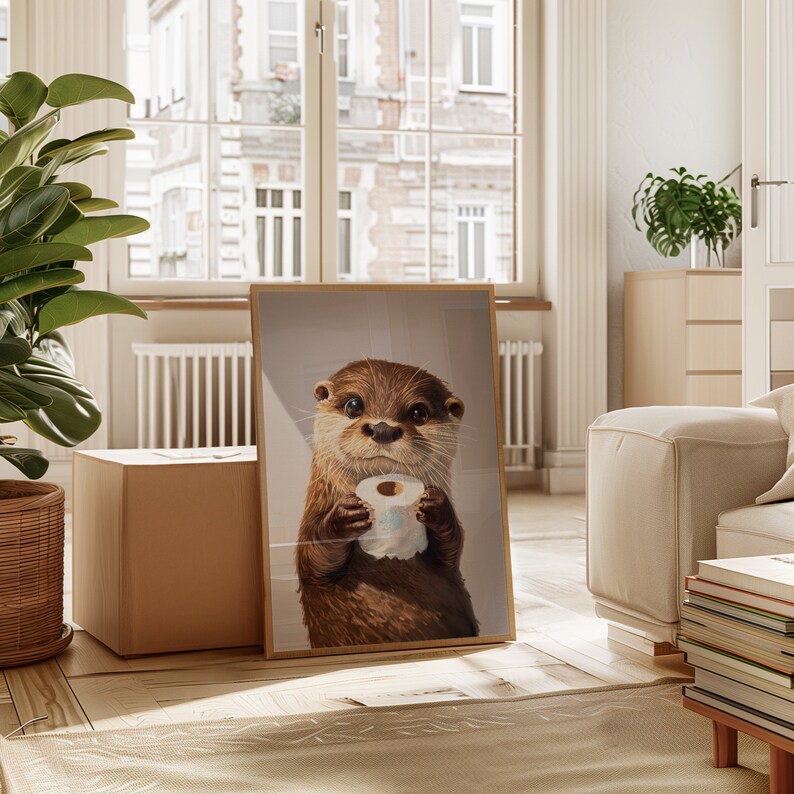 May include: A framed wall art print featuring a realistic illustration of a brown otter with dark eyes holding a small roll of white toilet paper in its paws. The otter is depicted from the chest up against a neutral, light-colored background.