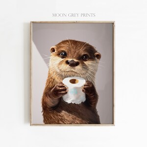 May include: A framed wall art print featuring a brown otter with large dark eyes holding a roll of white toilet paper. The text MOON GREY PRINTS is visible at the top of the print. The otter has detailed fur texture and whiskers against a light gray background.