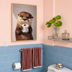 May include: A framed wall art print featuring a brown otter holding a roll of white toilet paper. The otter has dark eyes, a wet nose, and light brown whiskers. The print is displayed in a light wood frame against a light pink wall.