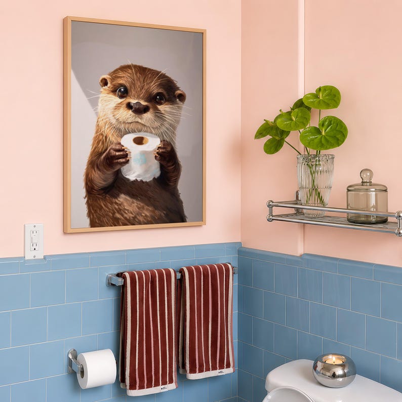 May include: A framed wall art print featuring a brown otter holding a roll of white toilet paper. The otter has dark eyes, a wet nose, and light brown whiskers. The print is displayed in a light wood frame against a light pink wall.