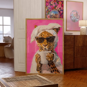 Op de afbeelding: Een ingelijst schilderij van een luipaard met een zonnebril en een witte handdoek op zijn kop, die een witte mok vasthoudt. De luipaard staat op een roze achtergrond.