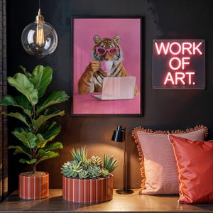 May include: A framed wall print featuring a tiger wearing pink glasses, drinking from a cup, and using a laptop against a pink background. Beside it, a neon sign reads WORK OF ART. Below, a fiddle leaf fig and a succulent planter sit on a surface.