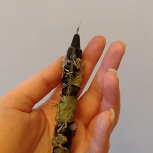 May include: A black candle with dried herbs and leaves wrapped around it. The candle has a white wick.