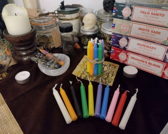 May include: A collection of colorful candles arranged on a dark brown surface. The candles are white, orange, black, green, yellow, blue, purple, pink, and red. There are also several boxes of incense sticks in the background, including Palo Santo, White Sage, Rosemary, and Dragon's Blood.