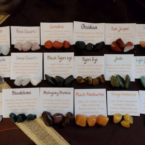 May include: A collection of 16 different types of crystals and stones, each with a small card describing their properties. The crystals are arranged in four rows of four, with each row featuring a different color scheme. The crystals are all small, ranging in size from about 1 to 2 inches.