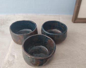 May include: Three small ceramic bowls with a dark blue glaze and brown accents. The bowls are arranged in a triangular shape on a light beige surface.