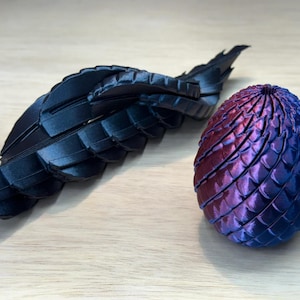 Puede incluir: Un objeto de ala articulada en negro mate colocado junto a un huevo ovalado con una superficie texturizada similar a escamas, que cambia de color de violeta intenso a azul metálico, con una pequeña abertura circular en la parte superior.