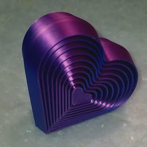 Pode incluir: Objeto em forma de coração multicamadas em tons de roxo e azul. O objeto, possivelmente impresso em 3D, apresenta contornos de coração aninhados. A cor muda de um roxo escuro para um azul brilhante, criando um efeito gradiente.