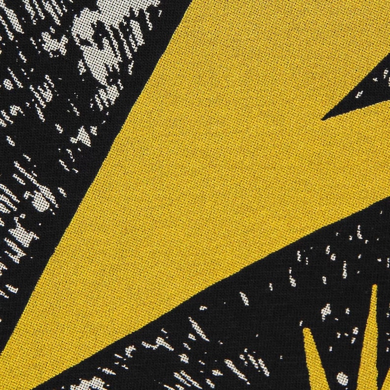 May include: A textile fabric featuring a bold geometric pattern with large, solid mustard yellow triangular shapes contrasted against black sections. The black areas are filled with a white, speckled, stippled texture that creates a grainy, distressed visual effect across the surface of the woven material.
