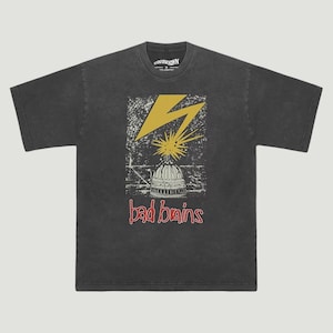 May include: A charcoal gray short-sleeve t-shirt featuring a graphic print of a yellow lightning bolt striking a dome structure. Below the graphic, the text "bad brains" is printed in a red, stylized font. The shirt has a crew neckline.